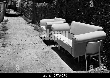 Photo noir et blanc d'un canapé abandonné et d'une chaise debout sur un chemin public à côté d'une haie, aucune personne n'est visible Banque D'Images