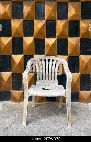 Gros plan d'une ancienne chaise en plastique blanc sale et cassée debout sur un sol devant un or et mur à carreaux noirs d'un établissement fermé Banque D'Images