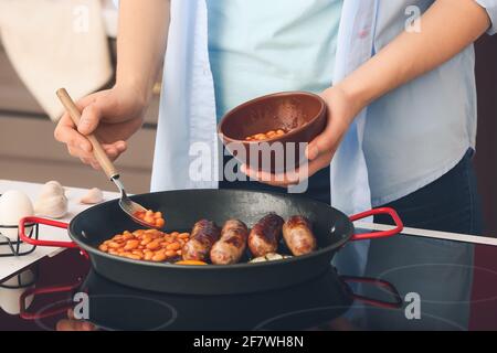 Femme qui cuisine le petit déjeuner anglais traditionnel sur cuisinière électrique dans la cuisine Banque D'Images