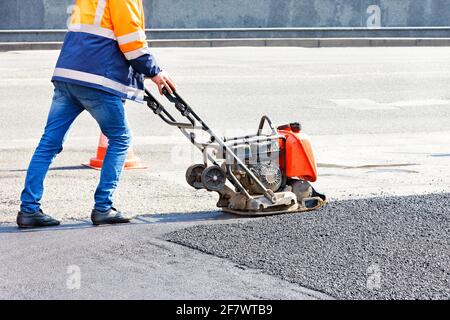 Un employé de l'entretien routier répare une section de la route par temps ensoleillé, compactant de l'asphalte frais avec une plaque vibrante. Copier l'espace. Banque D'Images