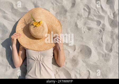 Une femme méconnaissable se trouvant sur le sable et recouvrant son visage d'un gros chapeau de paille. Vue d'en haut. Banque D'Images