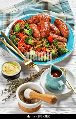 Saucisses de porc italiennes braisées avec des lentilles brunes, du poivre rouge et du brocoli sur une assiette bleue sur un fond de bois blanc avec des couverts dorés, de la moutarde, Banque D'Images