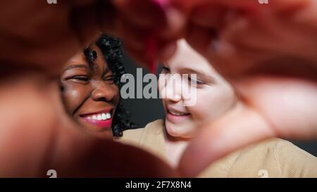 Belle-mère noire aux cheveux bouclés et blonde adopté école fille spectacle le cœur signe avec les mains et regarder dans les yeux avec joie sourit en gros plan Banque D'Images