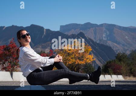 Jeune femme à capuchon blanc et lunettes de soleil sur fond gris pare-route en asphalte contre les arbres rouges et orange et rocailleux montagnes le jour de l'automne Banque D'Images