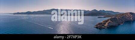 Vue panoramique aérienne du bateau passant par le phare de Revellata sur un promontoire rocailleux avec la citadelle de Calvi et le plafond de neige montagnes au loin Banque D'Images