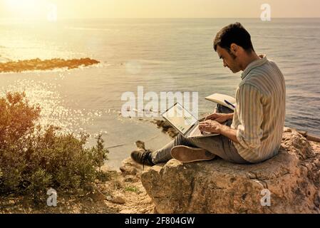 Un jeune homme utilise son ordinateur portable et son bloc-notes tout en travaillant à distance avec la mer en arrière-plan comme le soleil ensembles Banque D'Images