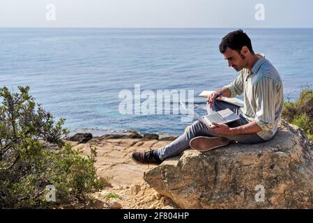 Un jeune homme utilise son ordinateur portable et son bloc-notes tout en travaillant à distance avec la mer en arrière-plan Banque D'Images