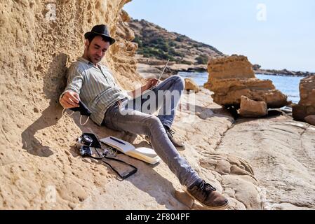 Un jeune entrepreneur télétravaille avec son ordinateur portable de la plage, laisse son masque facial à côté de son appareil photo, son smartphone et son bloc-notes Banque D'Images