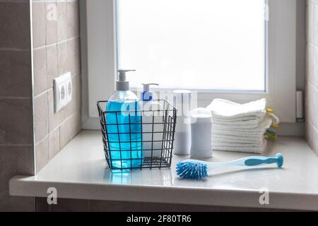 Une bouteille de lotion pour le corps utilisée, du savon liquide bleu se trouve sur un rebord de fenêtre dans une salle de bains avec carreaux beiges. Il y a de petites serviettes de toilette blanches en éponge dans une petite bla Banque D'Images