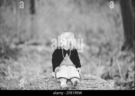 Une photo en niveaux de gris d'une petite fille blonde assise sur un rocher et un livre Banque D'Images