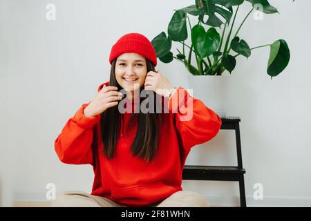 Sourit radieusement une femme dans un bonnet rouge vif et un sweat à capuche assis sur le sol avec ses jambes croisées. Elle a de longs cheveux droits sur les côtés. À l'intérieur, en fron Banque D'Images