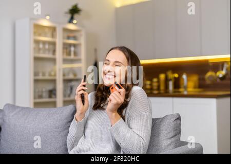 Photo d'une jeune femme excitée qui fait des achats en ligne tant attendus, heureuse femme tient un smartphone et une carte de crédit, magasiner en ligne, rêver avec les yeux fermés au sujet de nouvelles choses. Concept de consumérisme Banque D'Images