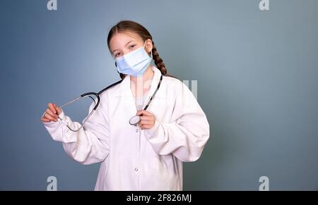 jolie jeune fille avec 2 tresses vêtues comme médecin avec masque de protection et stéthoscope devant un fond bleu Banque D'Images