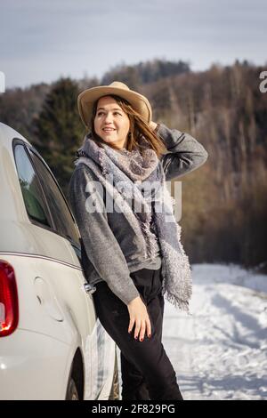 Femme avec un chapeau et une écharpe en plein soleil et se reposant à côté de la voiture pendant un voyage d'hiver. La jeune femme est à la recherche de paysage enneigé Banque D'Images