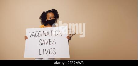 Fille portant un masque facial tenant une feuille avec "la vaccination sauve des vies" écrit. Enfant tenant une bannière sur fond marron. Banque D'Images