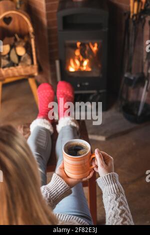 La femme boit du café chaud devant la cheminée et réchauffe ses pieds avec des chaussettes en laine. Femme se détendant dans le salon. Maison douce maison Banque D'Images