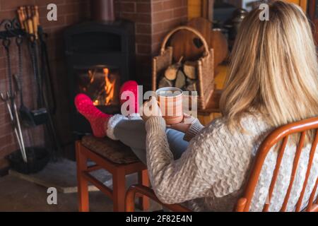 La femme boit du café chaud devant la cheminée et réchauffe ses pieds avec des chaussettes en laine. Femme se détendant dans le salon. Maison douce maison Banque D'Images