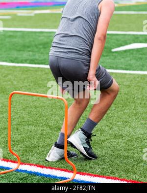 Un jeune garçon au camp d'été de football débarque après avoir sauté sur un obstacle en plastique orange sur un terrain de gazon vert. Banque D'Images