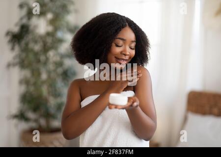 Jeune femme noire enveloppée d'une serviette appliquant de la lotion pour le corps ou crème après la douche à la maison Banque D'Images