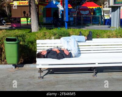 UKRAINE, YALTA - 29 JUILLET 2015: Homme sans domicile en jeans et veste noire dormant sur un banc blanc près de la poubelle verte dans la rue. Banque D'Images
