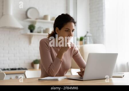 Jeune femme latina attentive regarder le séminaire en ligne sur ordinateur portable dans la cuisine Banque D'Images