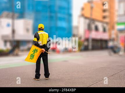 Vue arrière d'un homme japonais âgé portant un drapeau de passage de route « petit » imprimé et portant une casquette jaune, un gilet de sécurité, un brassard qui attend les enfants au ped Banque D'Images