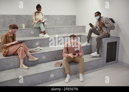 Grand angle de vue de divers groupes d'étudiants portant un masque tout en vous relaxant dans le salon scolaire moderne avec une distance sociale Banque D'Images