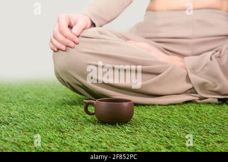 petite tasse de thé en argile vintage sur la moquette d'herbe à côté femme assise en position lotus sur fond de ciel voilé Banque D'Images