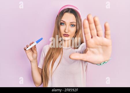 Jeune fille blonde tenant le résultat du test de grossesse avec main ouverte faisant signe d'arrêt avec une expression sérieuse et confiante, geste de défense Banque D'Images