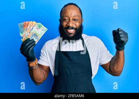 Un jeune afro-américain portant un tablier professionnel tenant des billets de banque en dollars australiens criant fier, célébrant la victoire et le succès très excite Banque D'Images
