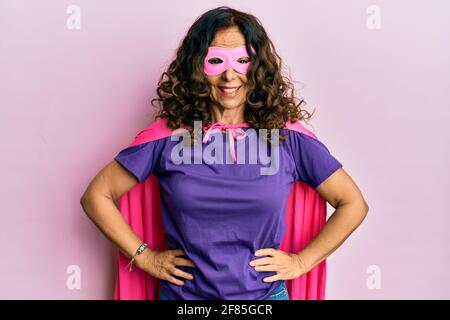 Femme hispanique d'âge moyen se déguquant en super-héros aux couleurs féministes rose et pourpre, souriant avec confiance Banque D'Images