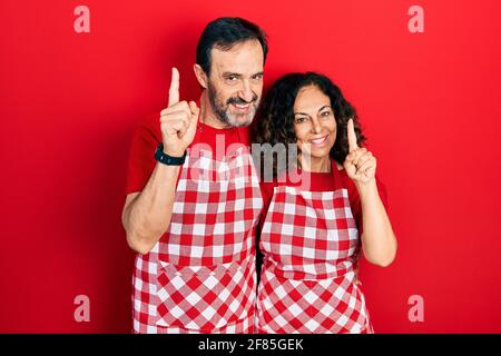Couple d'âge moyen de femme hispanique et homme portant un tablier de cuisinier pointant le doigt vers le haut avec une idée réussie. Quitté et heureux. Numéro un. Banque D'Images