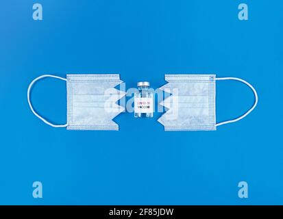 Flacon avec vaccin covid19 entre deux parties du masque médical découpé sur fond bleu. Banque D'Images