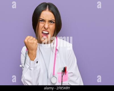 Belle femme hispanique portant l'uniforme du médecin et le stéthoscope en colère et fou élevant poing frustré et furieux tout en criant avec la colère. Rage et Banque D'Images