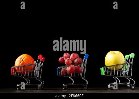 Close up, dark background image with three toy shopping carts filled with apple, tangerine and grapes A concept image for buying more healthy fruits a Banque D'Images