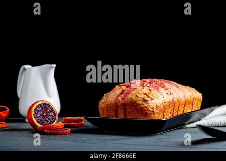 Image en gros plan d'un gâteau d'orange de sang frais fait maison dans un moule à pain et servi sur une assiette en porcelaine noire sur fond de pierre sombre. Il a l'orang de sang Banque D'Images