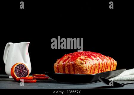 Image en gros plan d'un gâteau d'orange de sang frais fait maison dans un moule à pain et servi sur une assiette en porcelaine noire sur fond de pierre sombre. Il a l'orang de sang Banque D'Images