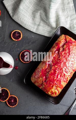 Image en gros plan d'un gâteau d'orange de sang frais fait maison dans un moule à pain et servi sur une assiette en porcelaine noire sur fond de pierre sombre. Il a l'orang de sang Banque D'Images