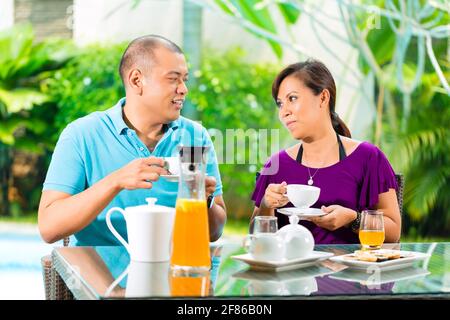 Asian couple having coffee sur le porche en face de leur maison, dans l'arrière-plan d'un jardin tropical Banque D'Images