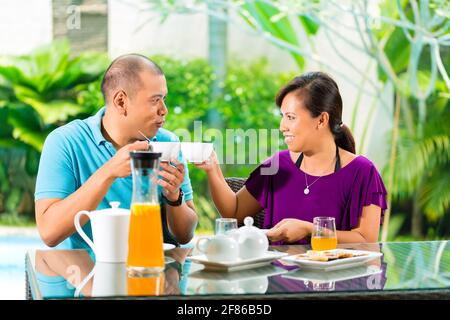 Asian couple having coffee sur le porche en face de leur maison, dans l'arrière-plan d'un jardin tropical Banque D'Images
