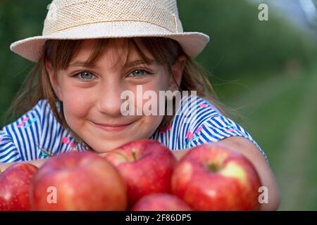 Petite fille souriante avec de beaux yeux regardant l'appareil photo. Concept d'alimentation saine. Gros plan de la petite fille avec chapeau et panier de pommes rouges. Banque D'Images