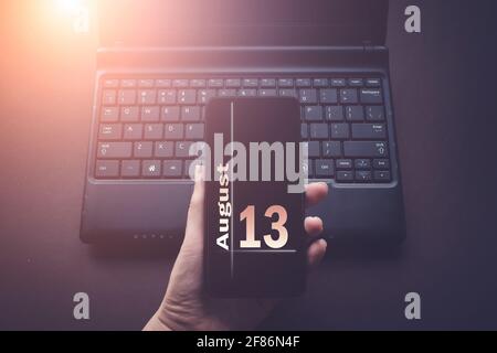 13 août. Jour 13 du mois, date du calendrier. Main tenant le téléphone mobile sur ordinateur portable arrière-plan sombre avec le soleil. Mois d'été, jour de l'année Banque D'Images