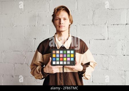 Jeune homme sérieux en tenue de travail tenant la palette carrée avec la couleur des échantillons tout en vous montrant contre la brique peinte en blanc mur Banque D'Images