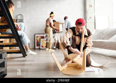 Jeune spécialiste de l'assemblage de meubles en vêtements de travail debout sur les genoux le plancher et l'assemblage de la nouvelle chaise en bois contre les colons Banque D'Images