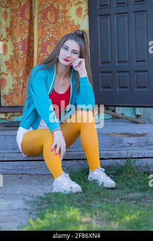 Une belle fille en vêtements lumineux se trouve sur le porche d'une maison de village. Femme dans le style des années 90. Banque D'Images