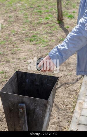 Une femme dans une veste bleue jette un verre jetable dans une poubelle de rue à côté du trottoir. Main d'une femme adulte avec une bague de mariage sur son tof Banque D'Images