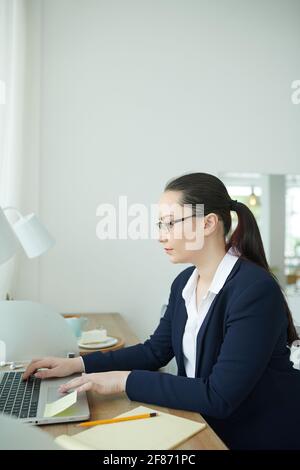 Jeune femme d'affaires sérieuse concentrée travaillant sur un ordinateur portable, vérifiant des documents, répondant à des e-mails et lisant des rapports Banque D'Images