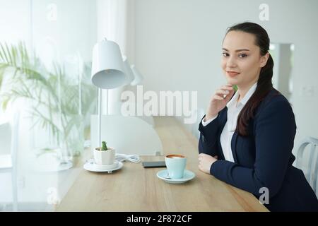 Portrait d'une jeune femme d'affaires souriante assise à table avec tasse de cappuccino et sourire à l'appareil photo Banque D'Images