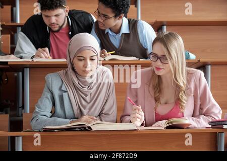 Deux étudiantes interculturelles de l'université assis à leur bureau salle de conférence et regarder à travers le passage dans le livre pendant le groupe travaillez en leçon Banque D'Images