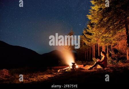 Vue magnifique sur le ciel étoilé de nuit sur une colline herbeuse avec des arbres et une femme voyageur près d'un feu de camp. Jeune randonneur assis près d'un feu de joie sous un beau ciel avec des étoiles. Banque D'Images